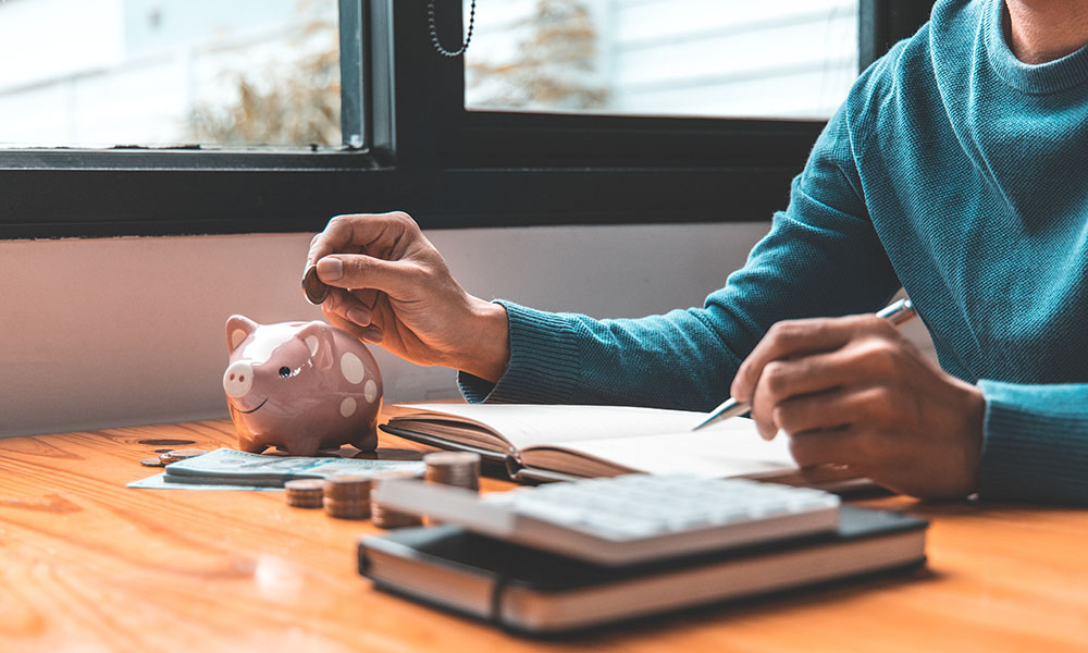 Una mujer inserta una moneda de un dólar en una hucha. En una libreta tiene apuntes del curso sobre finanzas para no financieros