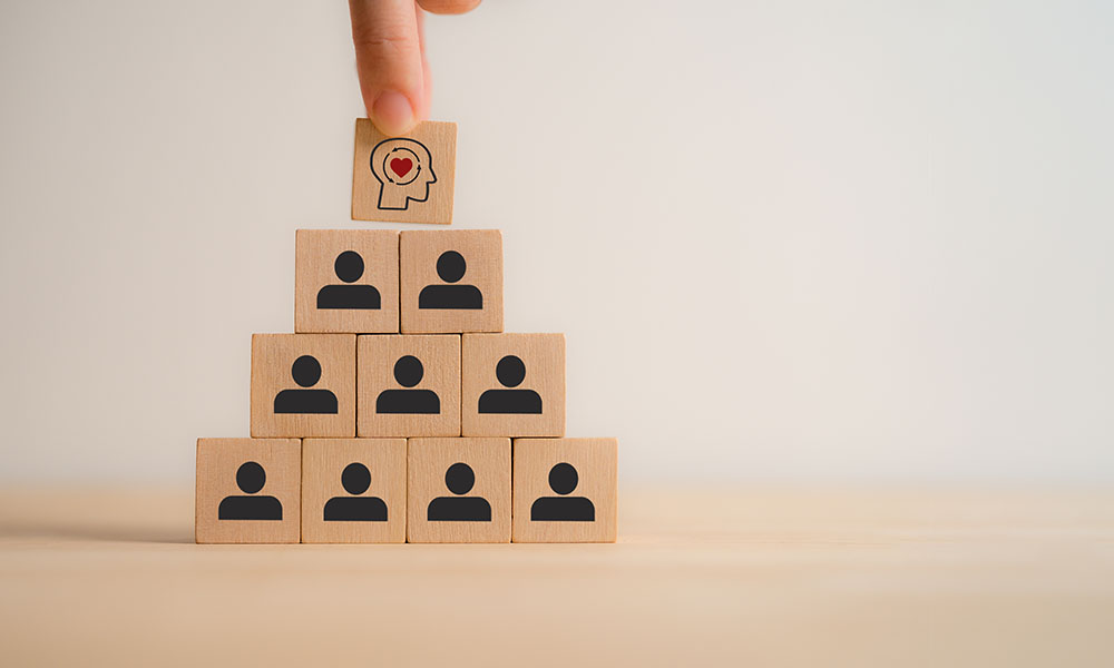 Bloques de madera apilados en forma de pirámide, donde una mano coloca en la cima un bloque con una cabeza y un corazón, simbolizando liderazgo, inteligencia emocional y gestión de equipos en el entorno empresarial